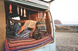 Ein Junge liest entspannt ein Buch, während er auf einer bunten Decke im offenen Heck eines Vintage-Campingbusses liegt, der am Meer geparkt ist.