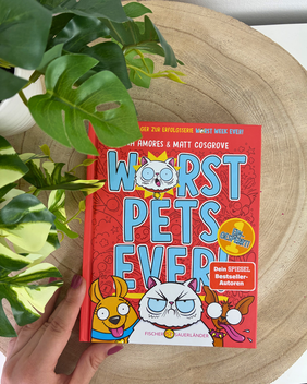 Eine Hand hält das rote Kinderbuch "Worst Pets Ever" von Eva Amores & Matt Cosgrove, das auf einer Holzscheibe neben einer grünen Pflanze liegt.