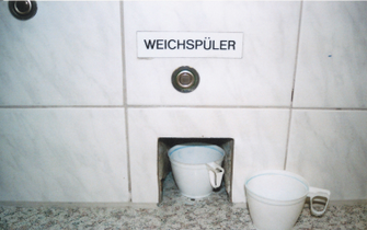 Ein Foto einer weiß gekachelten Wand, in einer Einbuchtung steht ein weißer Kaffebecher. Über der Einbuchtung steht: WEICHSPÜLER.