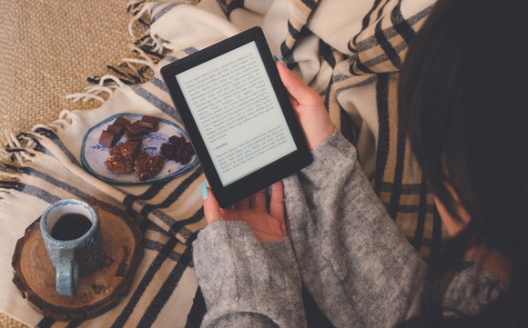 Eine Frau von oben fotografiert, während sie ein E-Book liest. Ein Kaffee und Plätzchen stehen nebendran.