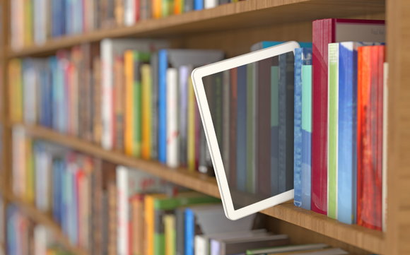 Foto von einem Bücherregal, in dem ein Tablet zwischen die Bücher gestellt wurde und zum großen Teil herausschau.t