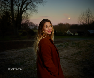 Porträtfoto von Miriam Georg, aufgenommen von Gaby Gerster. Sie steht im Freien bei Dämmerung, trägt einen rostbraunen Mantel und lächelt über die Schulter.