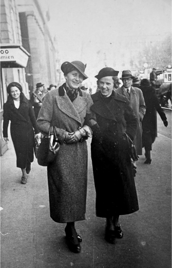 Schwarz-Weiß-Foto: Lina und Maria, Arm in Arm, lächeln in die Kamera. Sie tragen Mäntel und Hüte, im Hintergrund eine städtische Straße mit Passanten.