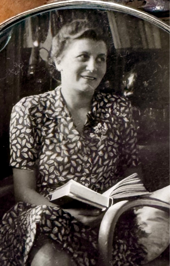 Schwarzweißfoto von Hedy. Sie lächelt und hält ein Buch im Schoß, während sie in einem Sessel sitzt. Blattgemustertes Kleid.