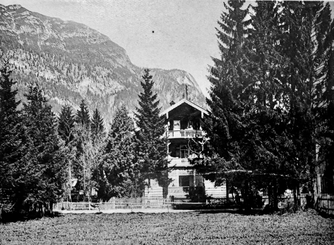 Schwarzweißfoto: Hotel Amalie in Garmisch-Partenkirchen. Holzhaus mit zwei Balkonen, umgeben von Tannen, vor Bergkulisse. Holzzaun im Vordergrund.