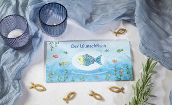 Das Bild zeigt das Wunschfisch Geldgeschenk vom Pattloch Verlag in einem passenden Ambiente mit kleinen Dekofischen und einem blauen Dekoschal