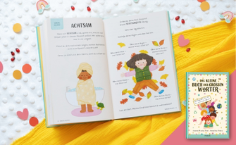 Das Bild zeigt das aufgeschlagene Buch "Das kleine Buch der großen Wörter" auf einem weißen Untergrund mit gelben Stoff. Um das Buch herum sind kleine Herzen und Regenbögen verteilt.