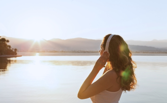 Eine Frau mit Kopfhörern lauscht Musik bei Sonnenuntergang über einem Gewässer.