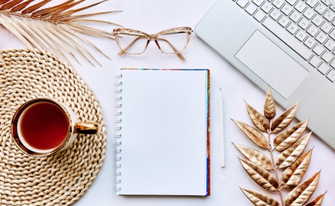 Draufsicht auf einen Schreibtisch mit Notizbuch, Stift, Laptop, Brille und goldfarbenen Dekoelementen.