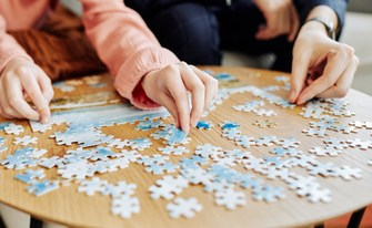 Ein weiß-blaues puzzle auf einem Holztisch, es sind zwei Personen an ihren Armen zu sehen. Sie puzzlen.