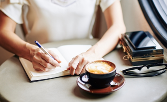 Eine Person sitzt an einem Tisch und schreibt in ein Notizbuch. Daneben stehen eine Tasse Cappuccino, eine Brille und ein Smartphone auf einer kleinen dekorativen Box.