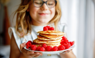 Nahaufnahme eines lächelnden Mädchens mit Brille, das einen Teller mit einem Stapel Pfannkuchen hält, die reichlich mit frischen Himbeeren garniert sind.