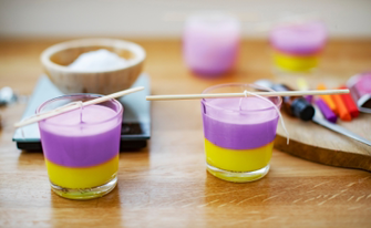 Nahaufnahme von zwei Gläsern, in denen zweifarbige Kerzen in leuchtendem Gelb und Violett gegossen wurden. Holzstäbchen halten die Dochte mittig fixiert, während das Wachs aushärtet. Im Hintergrund sind weitere Bastelmaterialien wie Farbpigmente und eine Waage zu sehen.