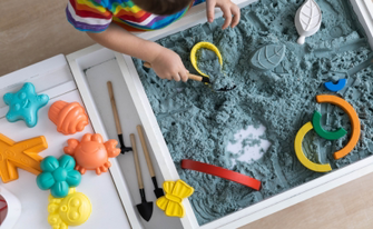 Draufsicht auf ein Kind, das in einem weißen Spieltisch mit blauem kinetischem Sand spielt. Verschiedene bunte Förmchen in Gestalt von Sternen, Krabben und Blumen sowie kleine Schaufeln liegen bereit.