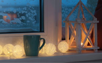 Nahaufnahme einer Fensterbank im Winter mit einer blauen Tasse, einer leuchtenden Kugel-Lichterkette und einer hölzernen Laterne mit brennender Kerze. Draußen ist Schnee am Fenster zu sehen.