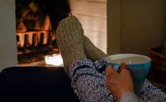 First-Person-Perspektive einer Person, die mit gemütlichen Wollsocken und einer Tasse Tee die Füße vor einem brennenden Kamin hochlegt.