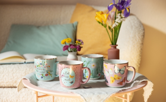 ier verschiedene Keramiktassen in Pastelltönen (Rosa und Hellblau) stehen auf einem kleinen Tisch. Die Tassen sind mit Goldrand, Blumenmotiven, Schmetterlingen und Texten wie „Kleine Auszeit“ oder „Zeit für mich“ verziert.