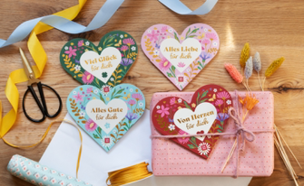 Vier herzförmige Geschenkbücher oder Karten sind dekorativ auf einem Holztisch arrangiert. Jedes Herz hat ein individuelles florales Design in frühlingshaften Farben und trägt einen positiven Schriftzug: „Viel Glück für dich“ (grün), „Alles Liebe für dich“ (hellrosa), „Alles Gute für dich“ (hellblau) und „Von Herzen für dich“ (dunkelrot). Die Szene wird ergänzt durch eine Schere, gelbe und blaue Dekobänder, ein in rosa Papier eingewickeltes Geschenk sowie getrocknete Blumen.