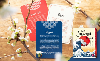 Eine Präsentation des Kartensets „Das Japan Orakel – Tägliche Weisheiten“. Auf einer hellen Holzoberfläche liegen drei Orakelkarten mit japanischen Lebensweisheiten: „Wabi-Sabi“ (rot), „Ikigai“ (weiß) und „Nagomi“ (dunkelblau). Die Karten enthalten erklärende Texte zu den jeweiligen Konzepten. Rechts unten ist die Verpackung des Sets abgebildet, die mit der berühmten „Großen Welle vor Kanagawa“ und der roten Sonne illustriert ist. Dekorative Kirschblütenzweige rahmen die Szene im Vordergrund ein.