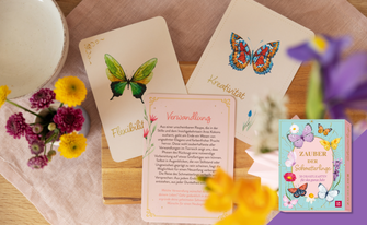 Eine helle, frühlingshafte Dekoration mit frischen Blumen und Orakelkarten, die Schmetterlinge sowie Begriffe wie „Flexibilität“, „Kreativität“ und „Verwandlung“ zeigen. Unten rechts ist die hellblaue Produktbox „Zauber der Schmetterlinge“ platziert.