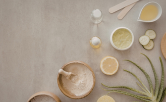 Draufsicht auf verschiedene natürliche Wellness- und Kosmetikzutaten, die auf einer grauen Steinfläche arrangiert sind. Zu sehen sind Holzschalen mit Pulver und Badesalz, eine Aloe-Vera-Pflanze, eine Zitronenscheibe, Gurkenscheiben, Öle in Fläschchen sowie Cremes und Holzspatel. Die linke Bildhälfte bietet freien Raum für Text.