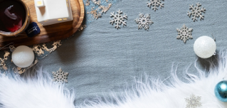 Ein gemütlicher, winterlicher Hintergrund in der Draufsicht auf einer graublauen Textiloberfläche, die viel freien Platz für Text bietet. Am oberen linken Rand steht ein Holztablett mit einer Tasse Tee und einem kleinen, weißen Deko-Häuschen. Der restliche Bereich ist weihnachtlich dekoriert mit silbernen Deko-Schneeflocken, weißen Glitzer-Weihnachtskugeln und einem kuscheligen, weißen Kunstfell-Besatz am unteren Bildrand.