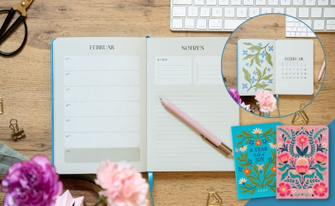 Aufgeschlagener Terminplaner 2027 auf Holzschreibtisch neben zwei Notizbüchern mit bunten Blumenstickereien.