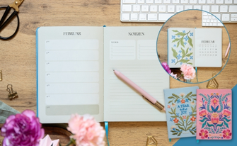 Aufgeschlagener Terminplaner auf Holzschreibtisch neben zwei Notizbüchern mit bunten Blumenstickereien.