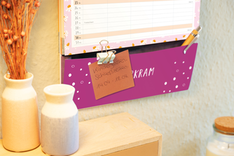Wandkalender mit lila Tasche für Notizen, festgeklemmtem Zettel, Kugelschreiber und Vasen auf Holzboard.