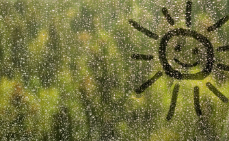 Regentropfen auf einer Glasscheibe, auf die eine lächelnde Sonne gezeichnet ist, mit unscharfem, grünem Hintergrund.
