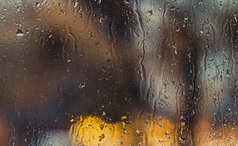 Regentropfen und Rinnen auf einer Glasscheibe mit verschwommenem, warmen Licht im unscharfen Hintergrund.