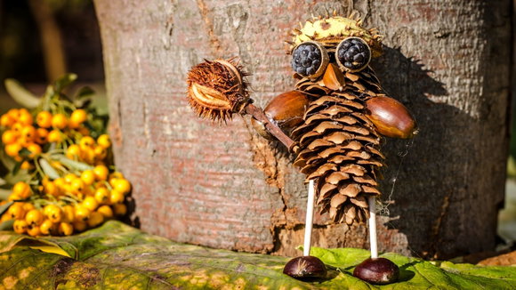 Gebasteltes Kastanienmännchen aus Tannenzapfen, Eicheln und Brombeeren vor einem Baumstamm im Herbst.