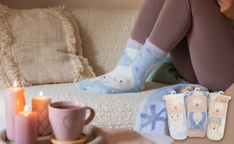 Eine Person in gemütlicher Kleidung sitzt auf einem Sofa und trägt hellblaue Kuschelsocken mit Tiermotiven. Im Vordergrund stehen brennende Kerzen und eine Tasse, rechts sind drei verpackte Sockenpaare zu sehen.