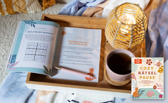 Ein hölzernes Tablett auf einer Decke, darauf ein geöffnetes Rätselbuch („Cozy Rätselpause“) mit Stift, eine Tasse Tee und eine leuchtende Korb-Tischlampe. Unten rechts ist das Buchcover eingeblendet.