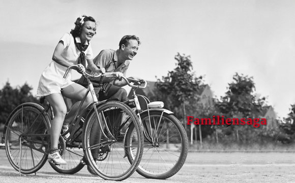Schwarz-weiß-Foto: Ein junges Paar fährt lachend Fahrrad, im Hintergrund ein Dorf.