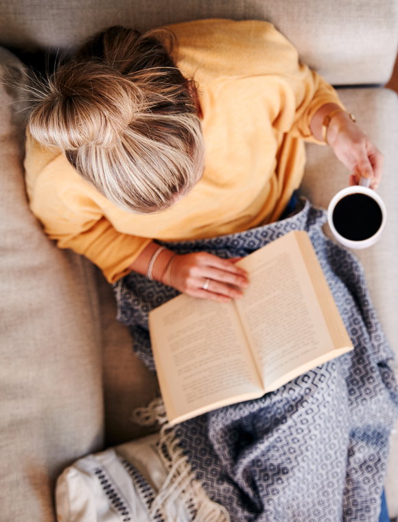 Foto von oben: Eine blonde Frau sitzt lesend auf dem Sofa, eine Wolldecke auf den Beinen, ein Kaffee in ihrer linken Hand