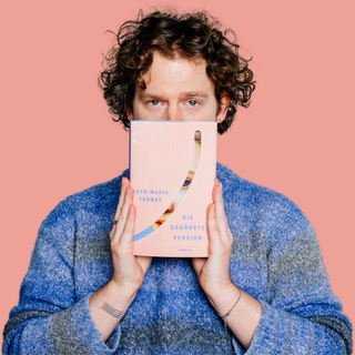 Ein Mann mit Locken und einem Flanellhemd hält «Die schönste Version» von Ruth-Maria Thomas vor den Mund