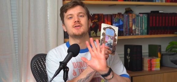 Ein junger Mann in einem Dungeons-and-Dragons-T-Shirt sitzt blickt zu uns (also der Kamera) hinter sich ein Bücherregal, vor sich ein Mikrofon, in der linken Hand hält er ein kleines Taschenbuch, die rechte Hand ist davor ausgestreckt.
